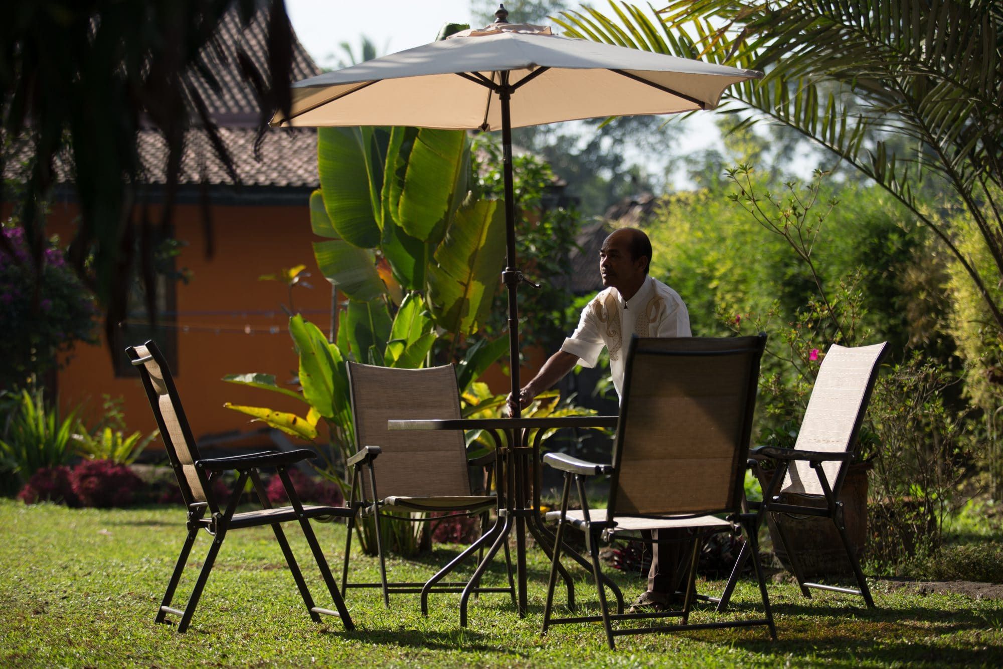 A member of Villa Tanen's staff setting a garden table with a parasol in the morning light, the house and tropical garden behind